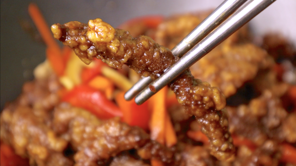 One piece of crispy beef in a chopstick with beef and bellpepper in the background