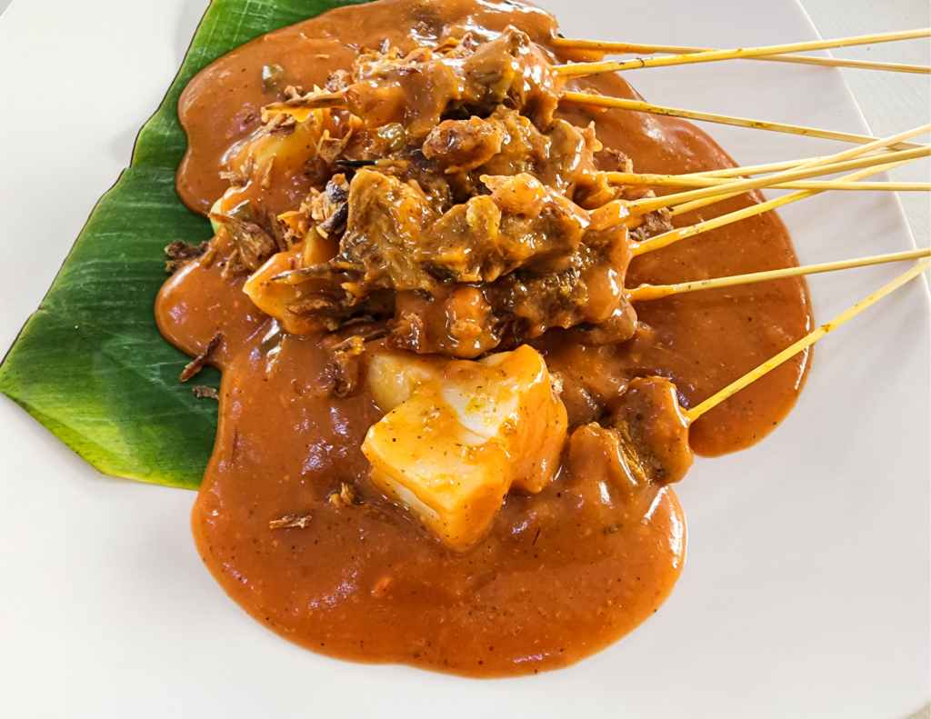 Indonesian satay with skewered meat served with peanut sauce, accompanied by rice cakes and garnished with crispy shallots, presented on a banana leaf.