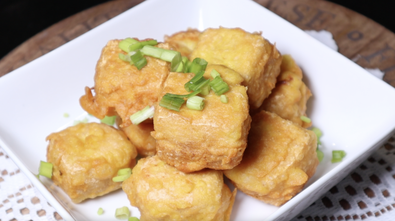 Close-up of crispy fried tofu cubes garnished with chopped green onions, served in a white bowl. The tofu is golden brown and crispy on the outside.