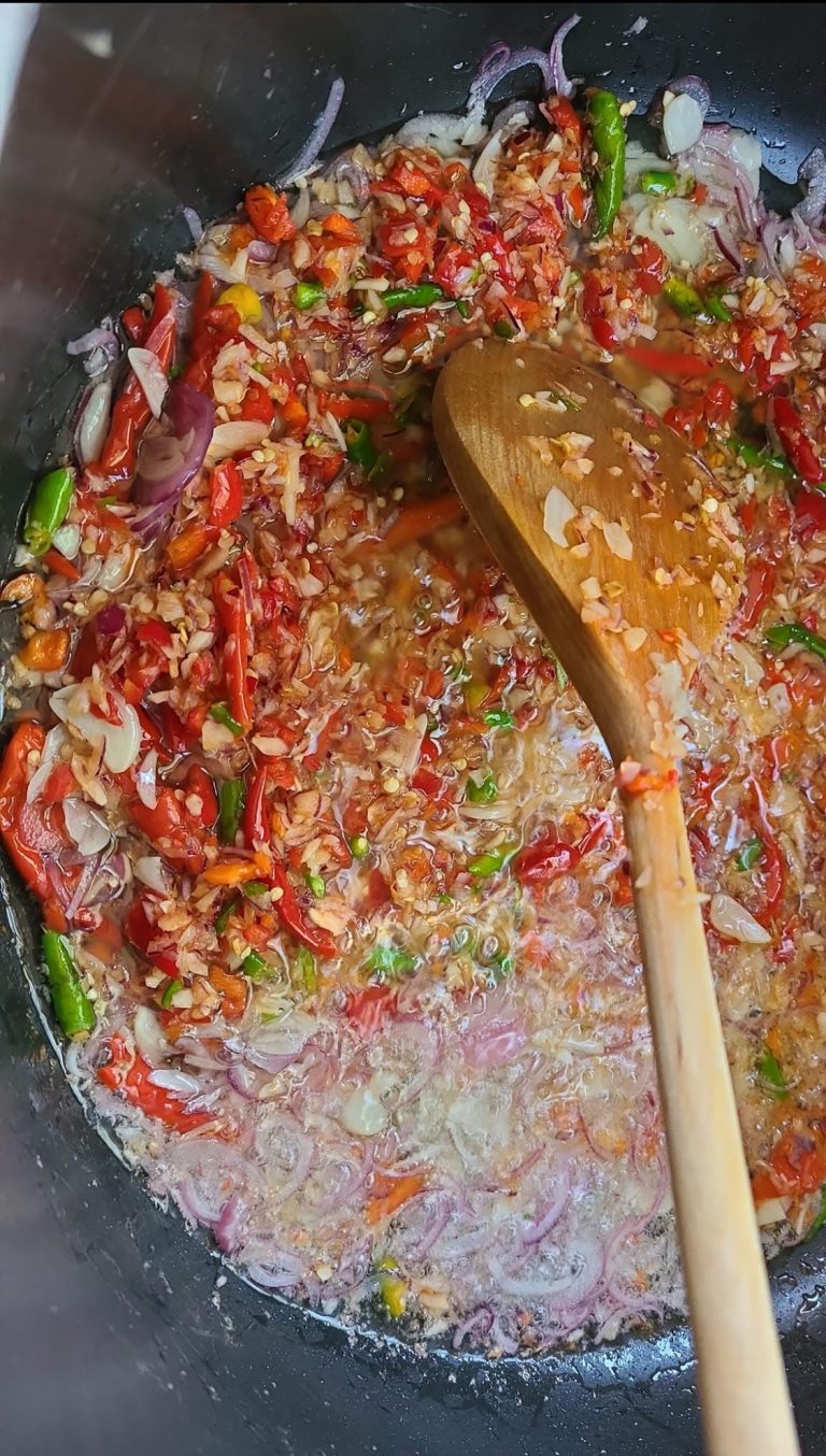 Close-up of a wooden spoon stirring a mixture of chopped red chilies, green chilies, and sliced shallots frying in a pan. The ingredients are sizzling in oil, with a few garlic cloves and bits of chili skin visible in the cooking process