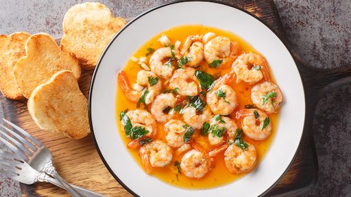 A plate of Spanish Garlic Shrimp (Gambas al Ajillo) served with toasted bread. The shrimp are cooked in a golden, garlic-infused olive oil sauce, garnished with chopped parsley, and accompanied by crispy slices of bread for dipping. The dish is presented on a rustic wooden board with silverware, capturing the vibrant colors and textures of this classic Spanish tapa.